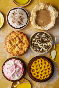 An overhead view of six pies with various toppings, including whipped cream, pink and white swirls, swirled hojicha and yellow squash rings, all sitting against a mottled yellow and white background.