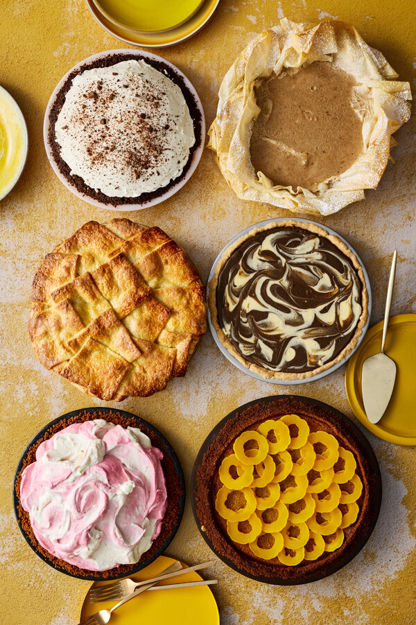 An overhead view of six pies with various toppings, including whipped cream, pink and white swirls, swirled hojicha and yellow squash rings, all sitting against a mottled yellow and white background.
